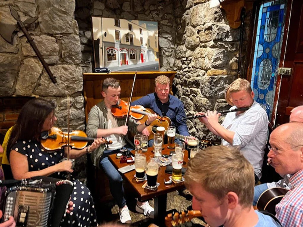 A group of people are playing traditional Irish music with fiddles, an accordion, and a flute around a small table with drinks in a cosy, stone-walled Ballinasloe pub.