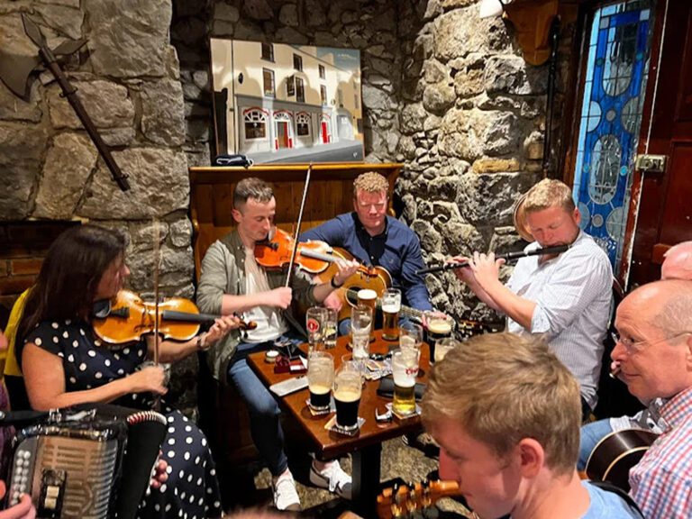 A group of people are playing traditional Irish music with fiddles, an accordion, and a flute around a small table with drinks in a cosy, stone-walled Ballinasloe pub.