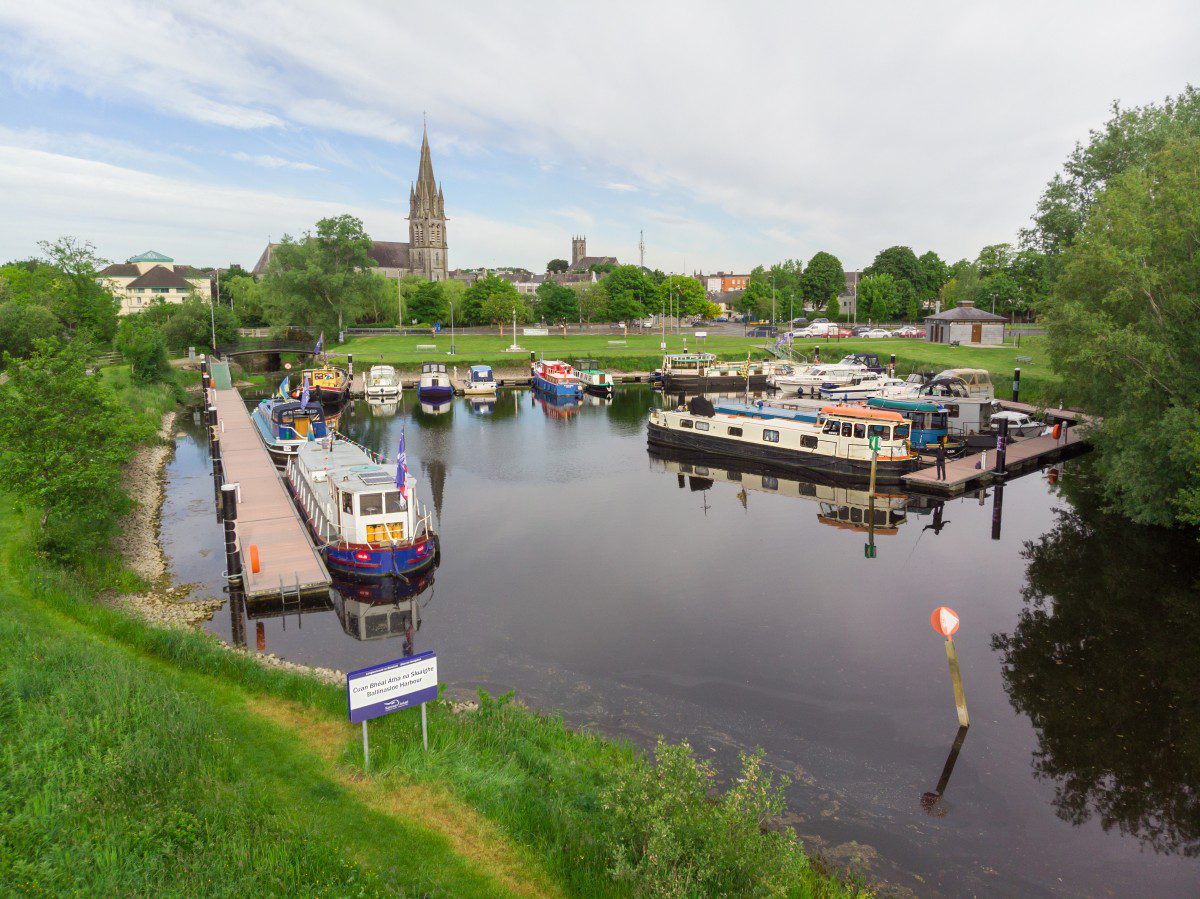 A small marina on a calm river in Ballinasloe, with several moored boats, green trees on both sides, a church with a tall spire in the background, and a grassy bank with a sign in the foreground.