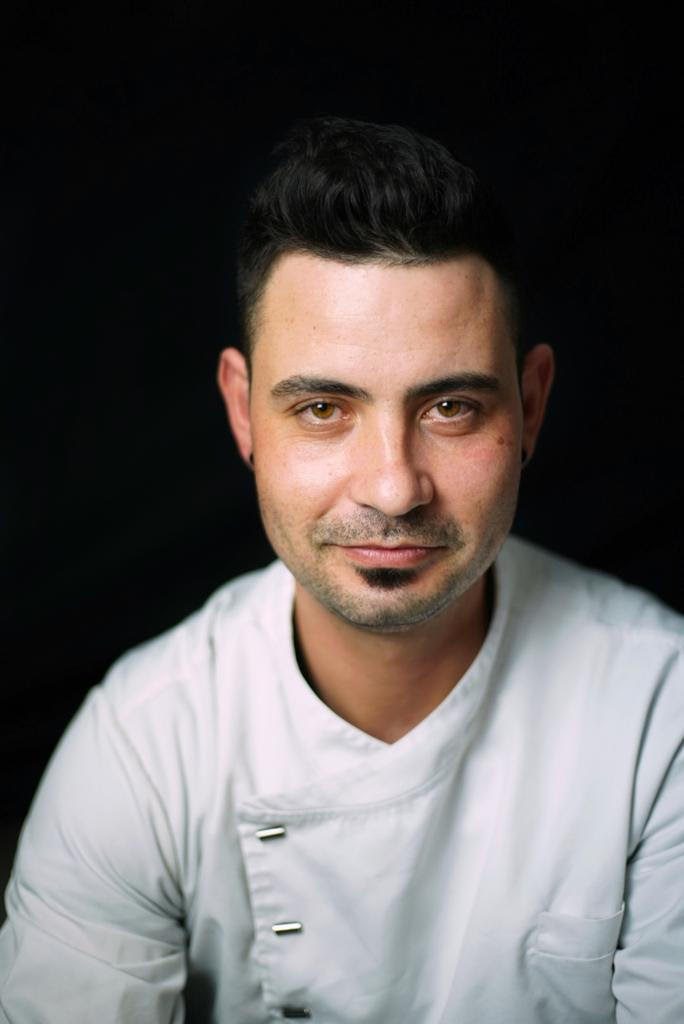 A man with short dark hair and facial hair, wearing a white chef’s jacket, smiles slightly as he poses against a black background—ready to lead the new team at the Golf Club Restaurant.