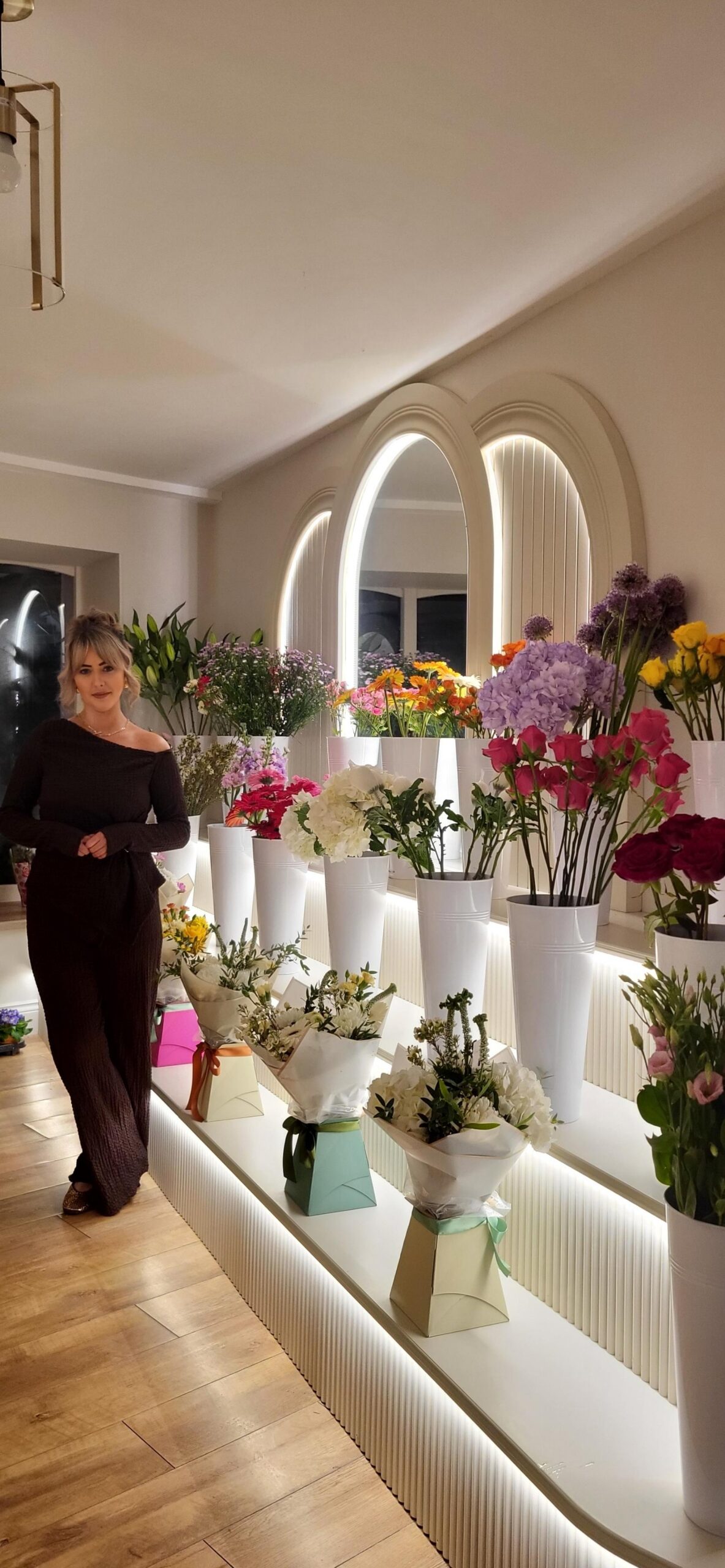 A woman in a dark dress stands indoors at Main St Florist, beside shelves filled with colourful bouquets and white vases of flowers, in front of two large, lit-up arched mirrors on the wall.