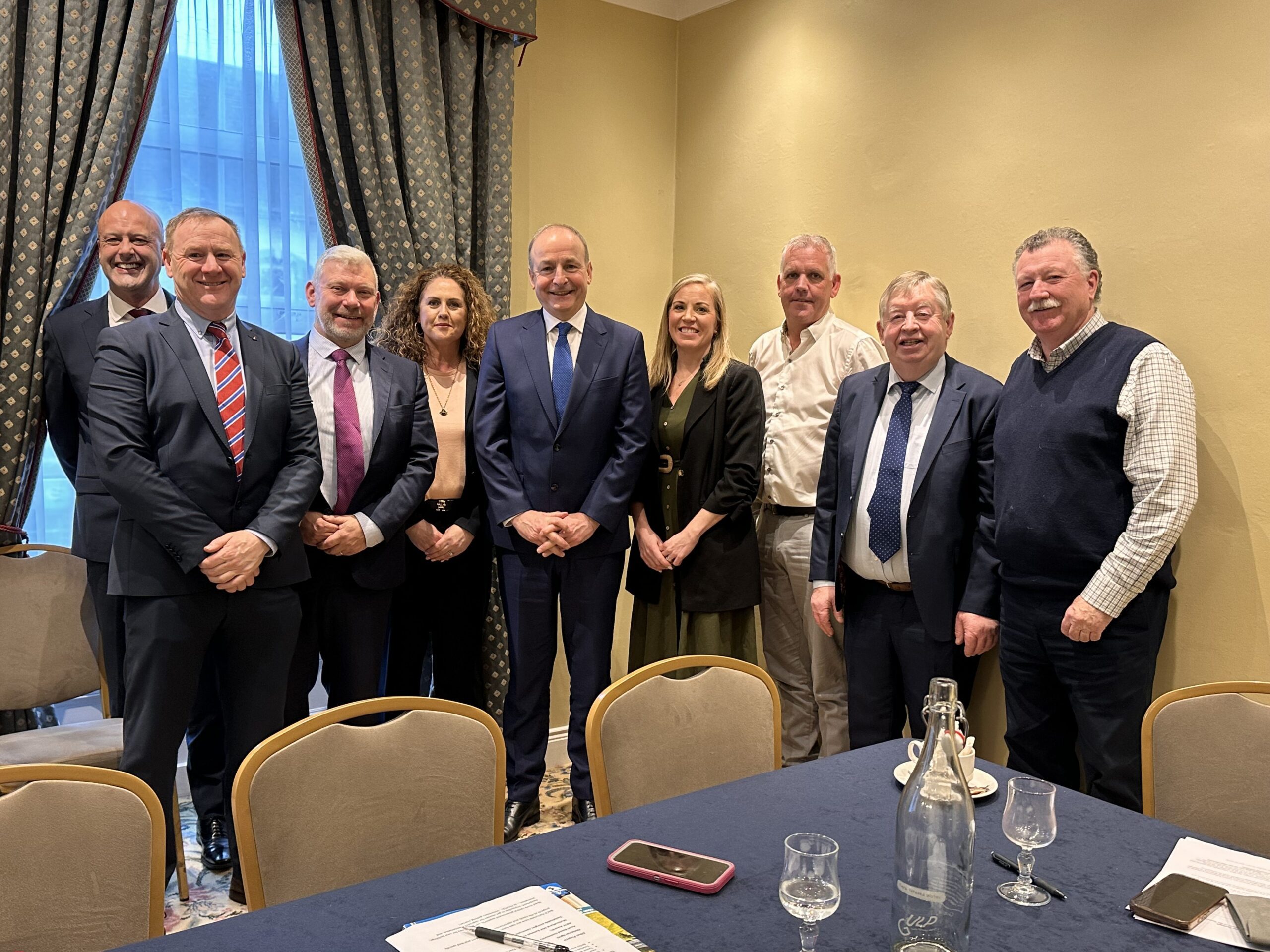 A group of ten people, dressed in business attire, stand and smile for a photo in a conference room with beige walls and patterned curtains. A table with papers, glasses, and a phone is in the foreground.