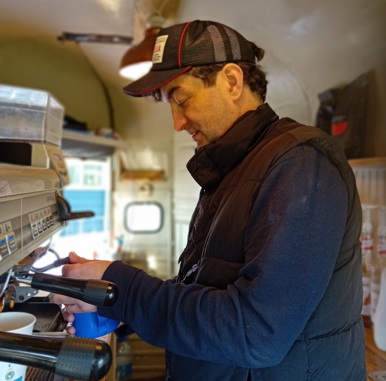 Ultan, Conlon, the owner of Wagtail coffee. Ultan is preparing a cup of coffee in his coffee truck. He's wearing a flat cap and a blue bodywarmer.