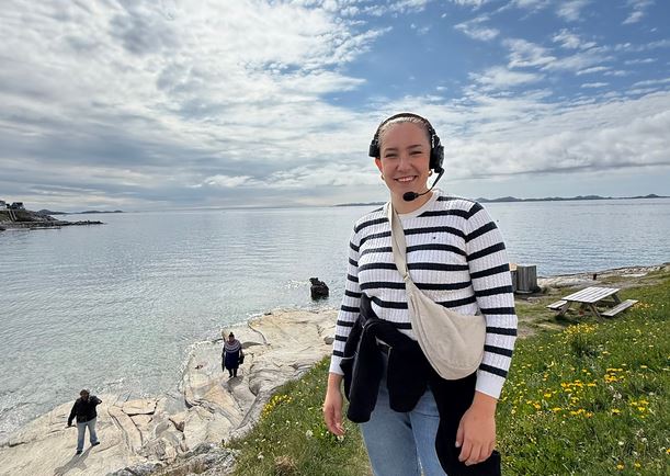 An award-winning TV Producer wearing a headset and striped jumper smiles by the sea. Behind her, two people stand on the rocky shore, and a picnic table sits on grassy ground with flowers. The sky is partly cloudy.