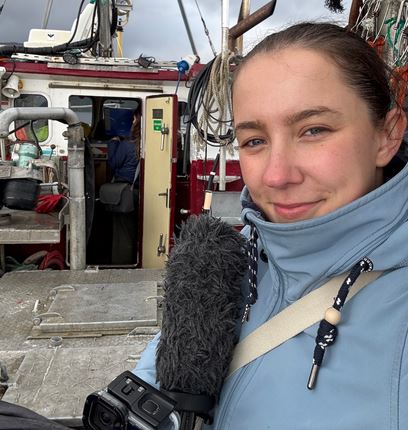 A woman in a blue jacket, an award-winning TV producer, holds a microphone and smiles at the camera while standing on a boat with equipment and ropes visible in the background.