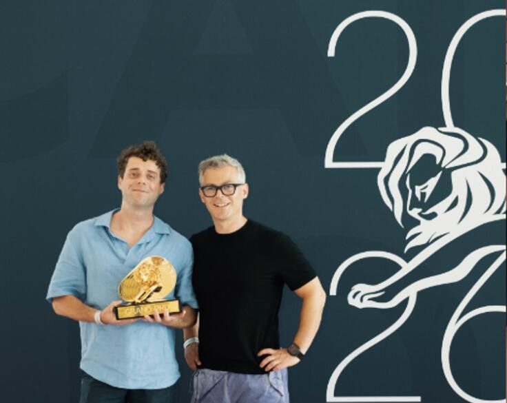 Two men from Ballinasloe stand in front of a blue backdrop with 2023 and a lion graphic. One holds a prestigious award, the golden GRAND PRIX lion trophy. Both are smiling and wear casual clothing.