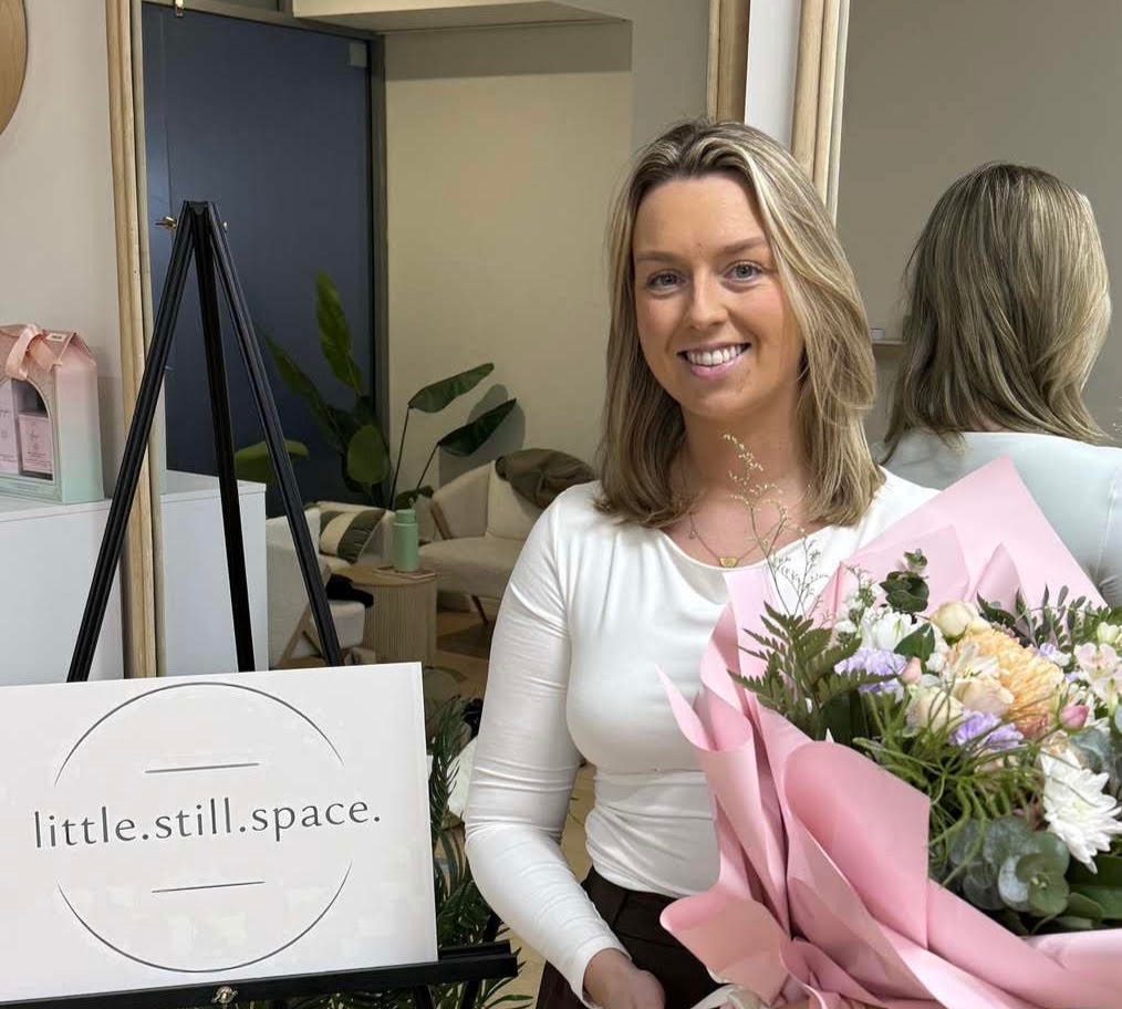 Michelle Jennings, founder of an app for children's wellbeing, Little Still Space. Michelle is pictured next to the little still space logo and is holding a bunch of flowers.