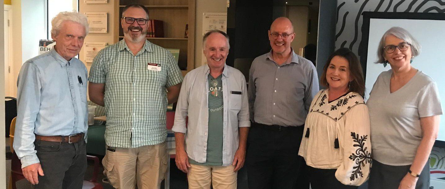 Six adults stand indoors, smiling for a group photo. Among them, five are men and one is a woman. Casually dressed, they appear to be in an office or meeting space—possibly at a local Toastmasters meeting—with shelves and a screen in the background. One person may be a visitor attending the gathering, adding to the welcoming atmosphere.