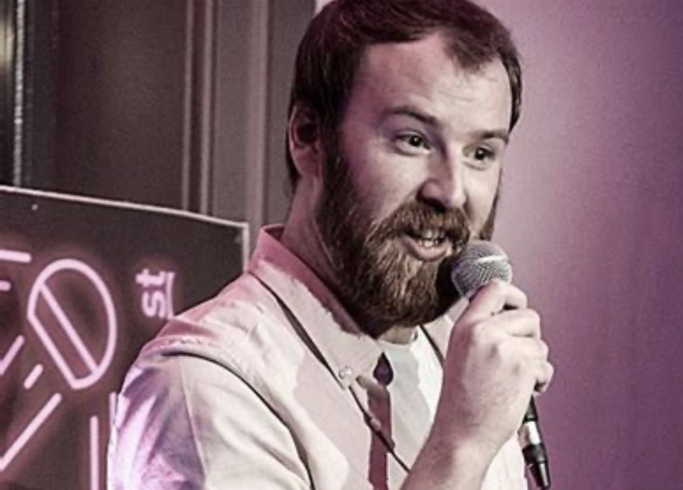 Comedian Danny Ryan. He is tall, with dark brown hair and a full beard and moustache. He's shown holding a microphone up to his lips.